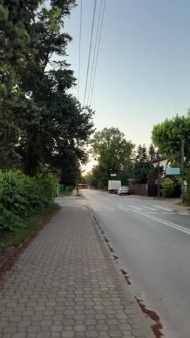 The video shows a street scene in Ożarów Mazowiecki, Poland, during what appears to be either dawn or dusk. The sun is low in the sky, casting a warm glow on the horizon. A paved sidewalk runs along the left side of the road, bordered by a lush green hedge and a large, dark green conifer tree. Overhead, several power lines stretch across the frame.

The street itself is paved and has a pedestrian crossing marked with white stripes. A white box truck and a silver car are parked on the right side of the road near the crossing. Beyond the vehicles, houses with fences and trees are visible, typical of a suburban or semi-rural setting. The overall mood is peaceful and serene, suggesting a quiet time of day. There are no people visible in the frame, and the primary subjects are the street, sidewalk, trees, and vehicles. The time of day is indicated by the low sun and the soft light, and the location context is provided by the text overlay.