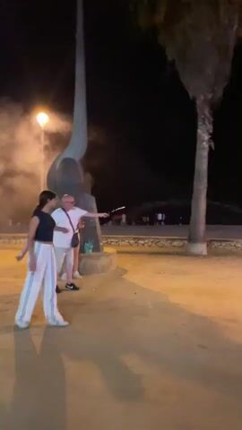 The video shows an outdoor scene at night. In the foreground, on the left, a woman in a white top and light-colored pants is standing. Next to her, a man in a white t-shirt and light-colored shorts is standing and holding a sparkler. He is looking to his right, toward the sparkler which is emitting sparks. Behind them, there is a large, abstract sculpture, possibly a bird or a sail, and a streetlamp. To the right of the man is a tall palm tree. In the background, there are other people and structures, and more sparklers are being lit, creating trails of sparks in the dark sky. The ground is sandy or dusty.