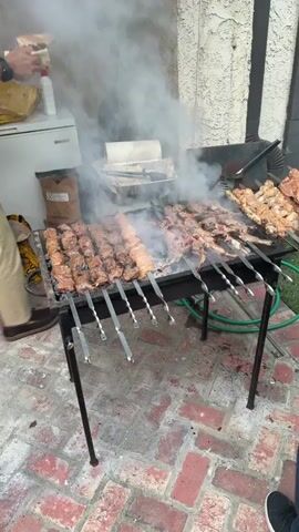 A close-up shot shows a rectangular barbecue grill with numerous skewers of meat cooking over hot coals, producing a lot of smoke. The meat on the skewers includes both smaller, cubed pieces and larger, flat cuts, possibly ribs or larger pieces of poultry. A person's arms are visible as they interact with the grill. In the background, there's a light-colored wall, and a green hose is visible on the ground near the grill.