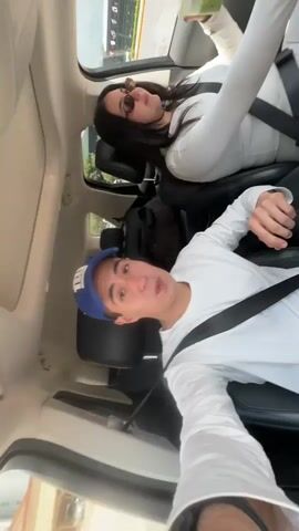 This is a video taken from inside a vehicle, appearing to be a selfie video, with two people visible. The person in the foreground, wearing a white long-sleeved shirt and a blue baseball cap, is looking at the camera and making a surprised or playful expression. A seatbelt is visible across their chest. Behind them, in the passenger seat, a person wearing a white long-sleeved outfit and sunglasses is also visible. This person is holding a green drink with what appears to be fruit in it and is looking away from the camera. Their seatbelt is also fastened. The interior of the car, including seats and part of the dashboard, is visible, as is the outside environment through the windows, suggesting the vehicle is in motion. The video is shaky, indicating movement.