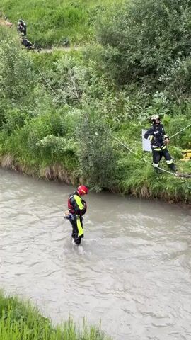 The video shows a car partially submerged in a river, tilted on its side. Several firefighters in yellow and black protective gear are present, some standing on the riverbank and others in the water. One firefighter is wading in the river, facing the car, while another is standing on a cable stretched across the riverbank, attending to the car. More firefighters are visible further up the bank, assisting with the operation. A red fire truck is parked on the road behind them. The environment is a green, grassy hillside with trees and vegetation. The river appears to be shallow in some areas. There are buildings and parked cars visible in the background, indicating a developed area. The overall scene suggests a rescue or recovery operation for the vehicle.