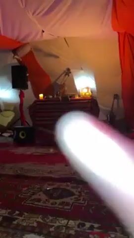 Inside a tent-like structure, the camera focuses on a table covered with a red and black striped cloth. On the table, there's a microphone stand, a musical instrument (possibly a violin), electronic equipment, a candle, and a decorative lamp. The floor is covered with a red patterned rug. A speaker and a guitar are visible on either side of the table. The tent walls are white and orange. A high-pitched sound can be heard.