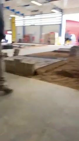 The video shows a group of people standing around a large pile of sand inside what appears to be a warehouse or large indoor space. The group consists of several individuals, some wearing casual clothing and others in uniforms. There is a large pile of sand in the center of the scene, with construction materials like planks and concrete blocks scattered around it. The background reveals warehouse doors and windows, as well as metal shelves and a red door. The group seems to be engaged in a conversation or observing something within the sand pit, as they are focused on the central area.