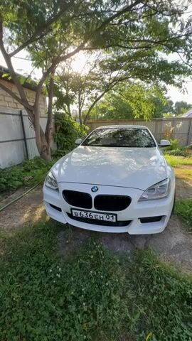A white BMW is parked in a yard. The car is facing the camera. Trees and a fence are in the background. The sun is shining through the trees.