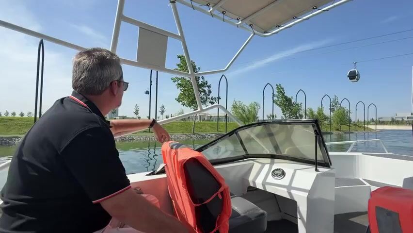 Two men are seen from a first-person perspective, riding in a white motorboat on a sunny day. The man in the foreground, wearing a black polo shirt and sunglasses, is seated on the left and points forward. An orange life vest is visible next to him. Another man, wearing a white t-shirt, is seated on the right. The boat has a windshield and a white canopy overhead. The water is calm, reflecting the clear blue sky.

The surroundings feature well-maintained green landscaping on the banks, with trees and decorative arch-like structures. In the distance, a modern, tall building, possibly a hotel or office, stands against the horizon. An aerial tramway (cable car) system spans the scene, with cabins visible along the cable. The overall impression is one of leisure, possibly a boat tour in an urban or resort area.