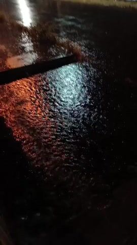 The video shows a street flooded with water at night. Rain is falling heavily, causing ripples and reflections on the water’s surface. Streetlights cast a warm glow on the wet pavement. A shadow, possibly of a barrier, is visible in the foreground. A building or wall runs along the side of the street. The overall scene is dark and wet.
