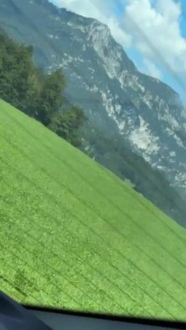 A video shows a view from inside a vehicle, looking out at a vast green grassy hill sloping upwards towards a majestic mountain range. The sky above is a clear blue with scattered white clouds. The grass on the hill appears to be meticulously mown, with visible lines indicating it was recently cut. Trees are sparsely scattered on the left side of the frame, clinging to the base of the mountains. The video captures the scenic beauty of a rural landscape, likely in a mountainous region.