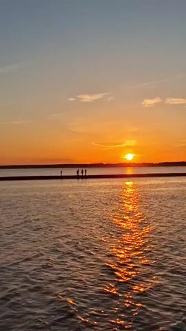 A sunset is shown over a body of water. The sun is low on the horizon, casting a golden glow across the sky and water. The sky is a gradient of orange, yellow, and blue. There are a few scattered clouds. In the middle ground, a dark line, possibly a pier or a landmass, stretches across the water. On this line, a small group of people can be seen. In the foreground, the water is rippled, reflecting the colors of the sunset. A small boat moves across the water from right to left in the distance.