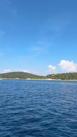 This is a landscape video shot from a boat on the water. The scene depicts a bright, sunny day with a clear blue sky dotted with fluffy white clouds. The sun appears to be high in the sky, suggesting it is late morning or early afternoon. The water is a deep blue and shows gentle ripples, indicating a calm sea.

In the background, a lush green island or coastline can be seen. The land is covered in dense vegetation, giving it a hilly appearance. There are some scattered structures visible on the land, and a few white sailboats are present in the distance, adding to the picturesque maritime setting. One sailboat is closer to the camera, with its white sail clearly visible against the blue sky. The overall mood is serene and tranquil, evoking a sense of peaceful vacation or leisure. The location is Mali Lošinj, Croatia, which is known for its beautiful islands and clear waters. There is no visible text in the video.