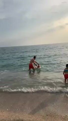 On a beach with tan sand, a man and two children are wading in the water. The man, wearing red shorts, is holding a young child. The second child is wearing red shorts and a striped life vest.  The water is turquoise with small waves coming to shore. The sky is light gray with a touch of sunlight peeking through the clouds.