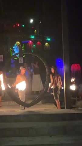 A man in a light-colored hat, white t-shirt, and beige shorts performs a fire show in front of a dark background with colorful lights. He is spinning inside a large, black, metal hoop that has two flaming torches attached to it. He is supported by another man in a black t-shirt and dark shorts, who is holding the hoop steady. A third man in a dark t-shirt is visible in the background on the left. The scene appears to be taking place at night.