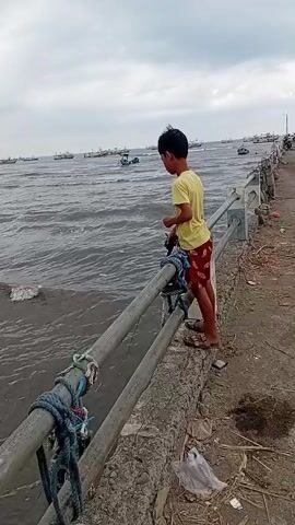 A boy in a yellow t-shirt and red shorts stands on a concrete walkway next to a railing, fishing. He is holding a fishing rod and appears to be casting his line into the water. The sea is visible with waves and several boats in the distance. The sky is overcast. The walkway is littered with some trash.