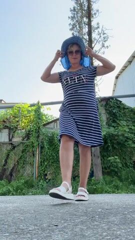 A woman with short, gray hair and sunglasses, wearing a navy and white striped dress and white sandals, poses outdoors. She holds a blue and white polka dot hat with both hands above her head at the beginning of the video and then lowers it. She then turns to her left and walks away from the camera. The scene is set outdoors with a concrete surface in the foreground, a fence and lush green foliage in the background, and a tall tree to the right of the woman. The sky is clear and blue.