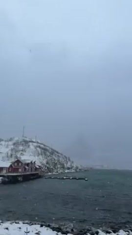 The video shows a coastal village in a snowy, overcast environment. Snow is falling. The water appears to be turquoise. There are several small islands and peninsulas with buildings, some of which are red. A snow-covered mountain is visible in the background, partially obscured by the heavy snowfall and fog. There are snow-covered rocks in the foreground. The camera pans slowly to the right.