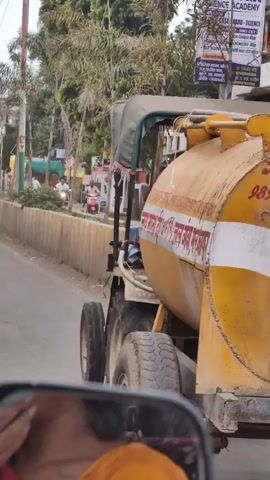 The video shows a blue tractor with a large yellow water tank attached, driving on a road. A man wearing a green shirt is driving the tractor. The tractor has a canvas canopy over the driver's seat. The water tank has text written on it in Hindi. In the background, there are buildings, trees, and power lines. A person on a red scooter can be seen in the distance. The video appears to have been filmed from inside another vehicle.