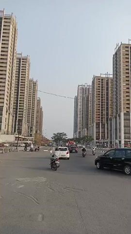 This is a vertical, eye-level, medium-shot video taken on a street with multiple tall buildings on both sides. The sky is visible above and appears overcast or hazy. The street is paved and appears to be a road with multiple lanes. There are various vehicles on the road, including cars, motorcycles, and scooters. Several people are riding on the motorcycles and scooters, some wearing helmets. The traffic appears to be moderate. The buildings are modern and appear to be apartment or office complexes, with many windows and balconies. There are also some trees visible in the distance, as well as streetlights and other urban infrastructure. The overall impression is of a busy urban environment.