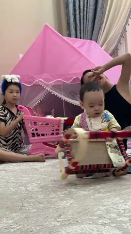 A woman is sitting on the floor in front of a pink tent. A young boy is also on the floor, playing with a toy cradle. The woman is smiling and making a peace sign with her fingers. Another young girl is sitting to the left of the boy, also making a peace sign. There is a pink toy shopping cart next to the girl. The scene is indoors, on a carpeted floor.