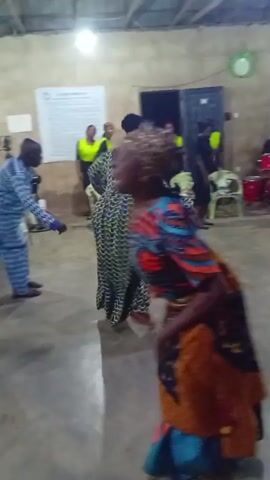 The video shows a group of people dancing in what appears to be an indoor hall or community center. The walls are plain and the floor is concrete. In the background, there are several chairs, a drum set, and a sign with the words "CAMP RULES" visible on it. 

Several individuals are dancing, primarily women, dressed in colorful traditional African attire. They move freely, extending their arms and swaying to the music, which is likely originating from the live band set up in the background. The atmosphere seems joyful and celebratory. A man in a blue and white patterned outfit is also dancing. Others are watching, including individuals wearing yellow safety vests.