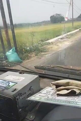 The video shows the view from the windshield of a vehicle driving past a rice paddy field. The field is green and stretches into the distance under a hazy sky. A white and red flag is planted on the side of the road. Electrical wires are visible overhead. Inside the vehicle, a car radio is mounted on the dashboard.