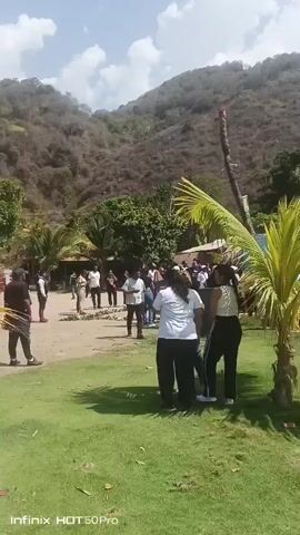 The video shows a group of people gathered outdoors in a tropical or semi-tropical setting. Most people are standing and appear to be conversing. The group is on a flat area covered with grass and sand. Behind them is a structure that could be a small building or a covered area. The background includes a mountain covered in foliage and a bright sky with some clouds. Palm trees are visible in the foreground. The video appears to have been taken with a mobile phone.