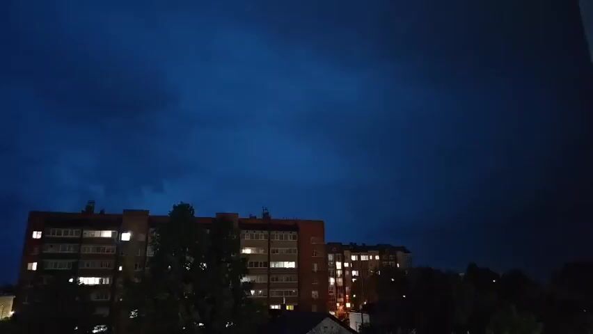 The video shows a dark, cloudy sky with lightning flashes over a residential area. Two multi-story apartment buildings are visible, with lights on in some windows. Trees are silhouetted against the sky between the buildings. The overall atmosphere is stormy and dark.