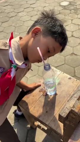 A young boy in a scout uniform is seen standing over a wooden table with a water bottle on it. The bottle is partially full and there is spilled water around it. The boy is looking down at the table. In the background, other children are visible, some in uniforms and some in casual clothing. The scene appears to be outdoors, possibly at a school or camp, with a paved ground surface made of hexagonal blocks. The lighting suggests it is daytime.
