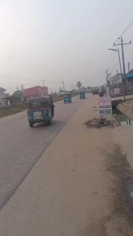 The video shows a road with vehicles moving on it. The view is from the side of the road. There are buildings and power lines visible in the background. A black car and a blue and white auto rickshaw pass by. Further up the road, there is a line of vehicles parked to the side. A sign with the words "Wash Available Here" is visible near the beginning of the video. The sky is overcast.