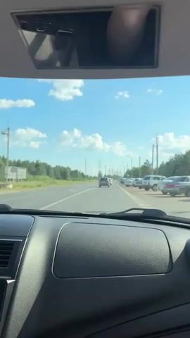 The video is taken from the interior of a car looking out through the windshield. The view shows a highway with a few cars ahead. On the right side of the road, there is a row of parked cars and trees. The sky is blue with scattered clouds. The car's dashboard and a sun visor are visible in the foreground. The sun visor is flipped down, and a hand is seen adjusting it. The video appears to be a recording of a car journey.