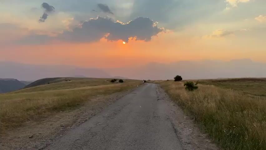 A paved road winds through a grassy meadow at sunset. The sun, partially obscured by clouds, casts a warm orange glow over the mountainous landscape. Two figures, a person and a dog, walk away from the camera on the road. The sky is a gradient of orange, pink, and purple, with rays of sunlight breaking through the clouds. The overall impression is one of peace and tranquility.
