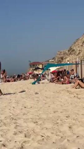 The video depicts a crowded beach on a sunny day. People are sunbathing on the sand, some under sunshades or tents. A building is visible behind the beach, near a cliff face. One man in white shorts walks in the foreground.