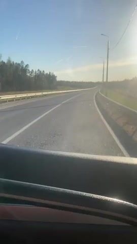 The video depicts a view from inside a vehicle driving on a two-lane road during the daytime. The road curves slightly to the right and is lined with guardrails on both sides. Streetlights are visible along the right side of the road. A forest lines the left side of the road. The sun is shining brightly, causing glare on the windshield. The interior of the vehicle is partially visible, including the top of a door panel.