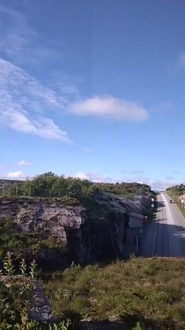 The video is a vertical shot taken from a grassy embankment, looking down and across a two-lane paved road that cuts through a rocky, sparsely vegetated landscape under a bright blue sky with scattered white clouds. The road stretches into the distance, appearing to ascend a gentle incline. On either side of the road, sheer rock faces rise, with some vegetation growing on top and at the base of the rocks. On the left side, a small, gray building with a red trim is visible nestled into the rock face, and some fencing is present. On the right side, there is more vegetation, including some trees and bushes, and a wire fence runs along the top of the embankment. The overall environment appears to be natural and somewhat rugged. There are no people visible in the video, and no actions are taking place beyond the implied movement of vehicles on the road, which are not clearly discernible.