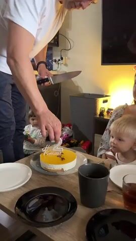 The video captures a birthday celebration. A man in a white t-shirt is holding a knife and appears to be about to cut a yellow cake with white frosting. The cake has a birthday candle in the shape of the number "2" and the words "Happy Birthday" written on it. Two young children and a woman are seated at a table. One child is looking at the cake, while the other is turned away from the camera. The woman is smiling and looking at the man. The environment appears to be a living room, with a television screen visible in the background. There are plates, cups, and a pitcher of tea on the table.