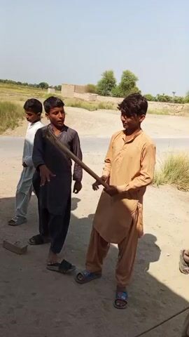 Two young males stand in a dirt area outdoors. One boy, wearing a dark grey shalwar kameez, stands facing forward and looks to the left. The other boy, wearing a light brown shalwar kameez, bends forward while holding a wooden stick. He is positioned to the right of the first boy. A third boy, wearing a white shalwar kameez, is partially visible on the far left of the frame. The background shows a dirt road, dry vegetation, and some buildings in the distance under a bright sky. The sun casts shadows on the ground.