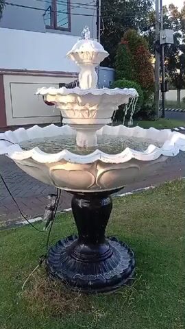 The video shows a three-tiered white fountain with a black base, situated on a patch of grass next to a paved walkway. Water flows from the top tier down to the middle and then to the large bottom basin. The fountain is decorated with ornate detailing. In the background, a building with a brown and white facade and a window is visible, along with trees and a street. Electrical cords are attached to the fountain. The video appears to be a close-up shot of the fountain, with no people or other animals present. The lighting suggests it is daytime.