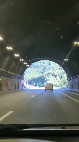 A white van drives out of a tunnel and onto a highway under a clear blue sky. The tunnel is dark and lit by overhead lights. The highway has multiple lanes marked with white lines and bordered by a metal guardrail. Trees and a mountain covered in lush green foliage are visible on the side of the highway. Several traffic signs are posted along the road, including signs with green arrows indicating lane directions. The view appears to be from the perspective of a driver inside a vehicle.