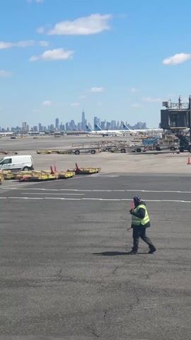 The video shows an airport tarmac under a sunny sky with a partial view of a cityscape in the background. The cityscape includes tall buildings, and what appears to be the New York City skyline. In the foreground, a person wearing a neon yellow safety vest, dark pants, and what appears to be a head covering walks across the asphalt tarmac. The person is holding a red object in their hand. Behind the person are several pieces of ground equipment, including a white van, various trailers, and some metal barriers. Further in the background are several planes, including ones with the "United" logo on them.