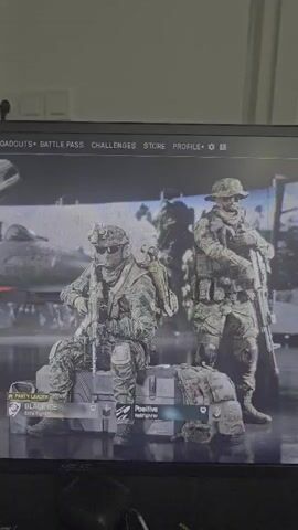 A screen displays the main menu of a video game. Two characters in camouflage military uniforms are visible in the foreground, standing in front of an aircraft. The character on the left is seated on a crate, while the character on the right is standing. Below the characters, player information and game menu options such as "LOADOUTS," "BATTLE PASS," "CHALLENGES," "STORE," and "PROFILE" are displayed. The overall scene is dimly lit, suggesting the game is being played in a low-light environment.