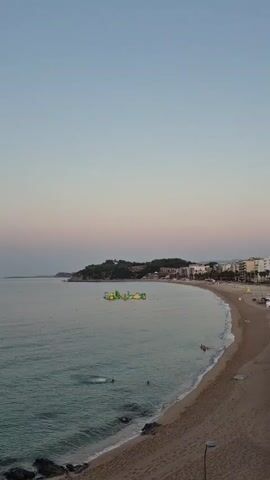 The video shows a calm ocean with a sandy beach in the foreground. The sky is a soft gradient of blue and pink, indicating either sunrise or sunset. On the water, there are several inflatable water attractions, possibly a water park, with people visible enjoying themselves. Further down the coast, there are buildings and trees. The overall scene is serene and suggests a vacation or leisure setting.