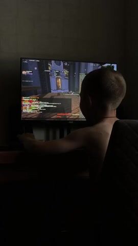 A young boy is playing a video game in a dimly lit room. He is sitting in front of a computer monitor, with his back to the camera. The monitor displays a colorful game scene with various characters and objects. The boy's right hand is on the mouse, and his left arm is resting on the desk. He is wearing a dark-colored garment draped over his shoulders. The room appears to be a bedroom, with a textured wall in the background. The overall atmosphere is one of concentration and immersion in the game.