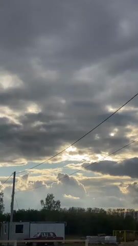 The video shows a view of a cloudy sky with sunlight breaking through the clouds. A utility pole with wires is visible in the foreground, along with trees and some structures in the background. There is a car parked near one of the structures, which appears to be a small building or portable unit. The ground is uneven and appears to be dirt or gravel, with some construction materials visible. The overall scene suggests an outdoor location, possibly rural or semi-rural, under an overcast sky.