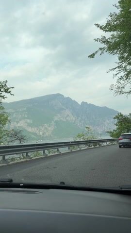 A car drives along a lakeside road toward the town of Valerana in what appears to be Italy. The road has a metal guardrail on the lake side and is lined by trees. On the other side of the lake is a steep, rugged mountain. The sky is cloudy. Music is playing in the car.