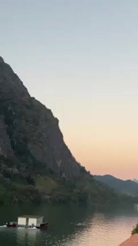 The video shows a tranquil scene with a large mountain towering over a wide river. The mountain is steep and rocky, covered in greenery. A small, white houseboat is docked on the river, and the river is calm with gentle ripples. The sky is a pale gradient of colors, transitioning from light blue to soft pink, suggesting either sunrise or sunset. The overall atmosphere is peaceful and serene.