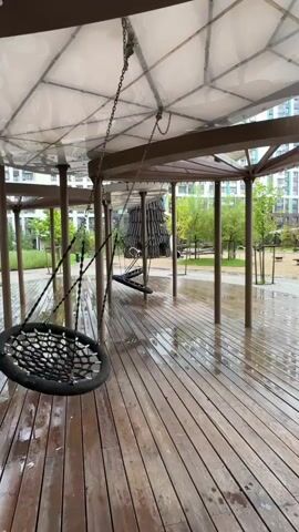 This is a video of a playground with several swings under a canopy. The ground is wet, indicating it has rained. The swings are made of netting and hang from chains. In the background, there are trees and buildings. The camera appears to be stationary.