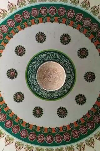 The video shows a circular ceiling of a building, decorated with colorful concentric circles. The innermost circle is green and surrounds a light fixture. The circle immediately outside of the green circle is white and contains a pattern of evenly spaced small flower designs. The next circle is decorated with alternating red and orange shapes, followed by an outer circle with repeating green and cream-colored motifs. The camera is pointed upwards.