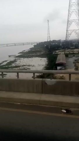 The video shows a wide river crossed by a long bridge, as viewed from a road on another bridge. The river is wide and calm, with a shoreline showing patches of waterlogged vegetation. On the near bank, there's a structure that seems like a dock or pier extending into the water, and some low buildings. A tall, thin communication tower stands on the shoreline, and another tower is seen in the right corner. The sky is overcast. The perspective is from a moving vehicle, likely crossing a bridge that is separate from the one in the background, as indicated by the presence of a roadside barrier with a gray railing.