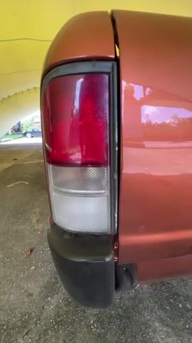 A low-angle, shaky, first-person perspective video begins with a close-up of the taillight of an orange pickup truck parked under a yellow, tent-like structure. The camera then pans to the right, revealing the side of the truck and a white pickup truck parked further ahead under the same structure. The video continues to pan right, moving closer to the white pickup truck, which is also parked under the yellow structure. The camera then focuses on the front of the white pickup truck as it moves forward slightly. The video ends with a view of the white pickup truck from the front, with trees and buildings visible in the background through the opening of the yellow structure. The ground appears to be concrete or asphalt and is covered with some debris.