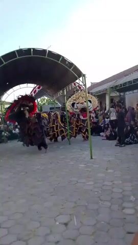 The video shows an outdoor cultural performance. In the foreground, a stone-paved area is visible. Two performers in elaborate costumes, one resembling a horse and another a lion-like creature, are engaged in a dance. The horse-like costume is black with red and white accents, and it has a large, feathery headdress. The lion-like costume is black and yellow, with a large, stylized head. A temporary canopy structure with a green roof and metal poles provides shade for the performers. In the background, a crowd of spectators, mostly seated, watches the performance. The environment appears to be a village or open-air venue, with buildings visible in the distance. The sky is clear and bright, suggesting it is daytime. The overall scene depicts a lively traditional performance with a cultural audience.