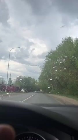 The video shows a view from inside a car driving on a wet road under a cloudy sky. Raindrops are visible on the windshield, and the wipers are running. On either side of the road are trees and roadside vegetation. The road ahead shows other vehicles, including cars and a truck, moving in both directions. In the foreground, the dashboard of the car and a portion of the steering wheel are visible. The speedometer shows that the car is moving.