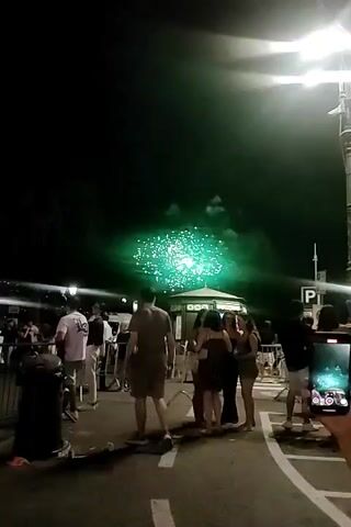 The video shows a street scene at night with fireworks in the sky. A group of people are gathered on the street, some are watching the fireworks, while others are talking or walking. There are streetlights and palm trees visible in the background. The fireworks are green and white, and they are bursting in the sky. The scene appears to be festive and celebratory.